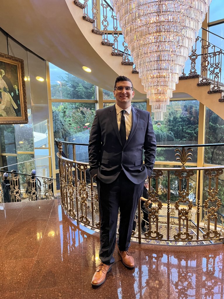 A man wearing glasses in a two piece suit infront of a chandelier leather brown shoes and smiling