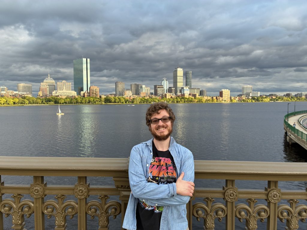 A guy wearing blue jean jacket with glasses infront of boston harbor smiling folded arms